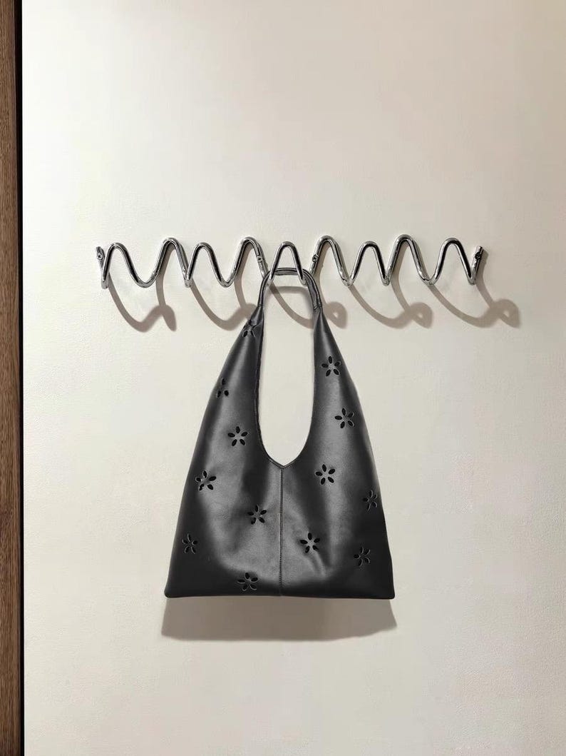 May include: A black tote bag with a floral cut-out design hangs from a wavy, chrome-colored metal rack. The bag has a single handle and a wide opening. The background is a plain, off-white wall.