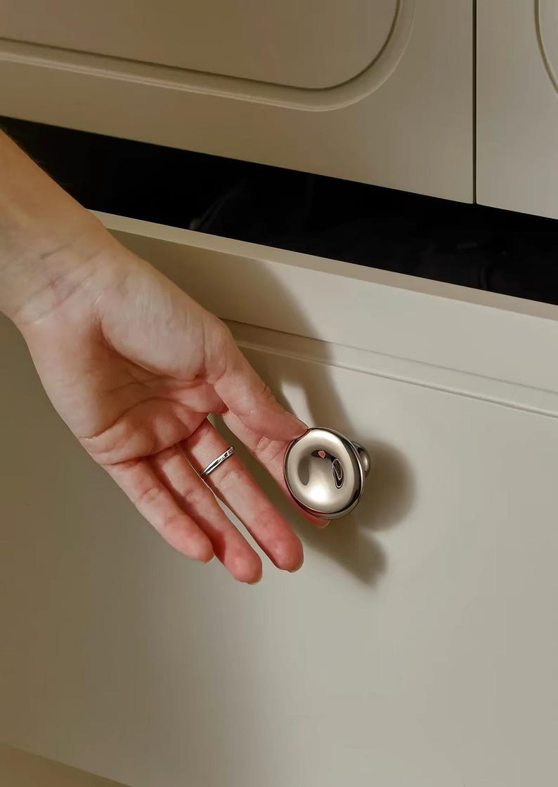 Puede incluir: Un pomo de armario circular, plateado y pulido, con una superficie lisa, reflectante y ligeramente abombada. El pomo est&aacute; montado en un frente de caj&oacute;n plano de color beige claro, con un dise&ntilde;o de bordes redondeados.