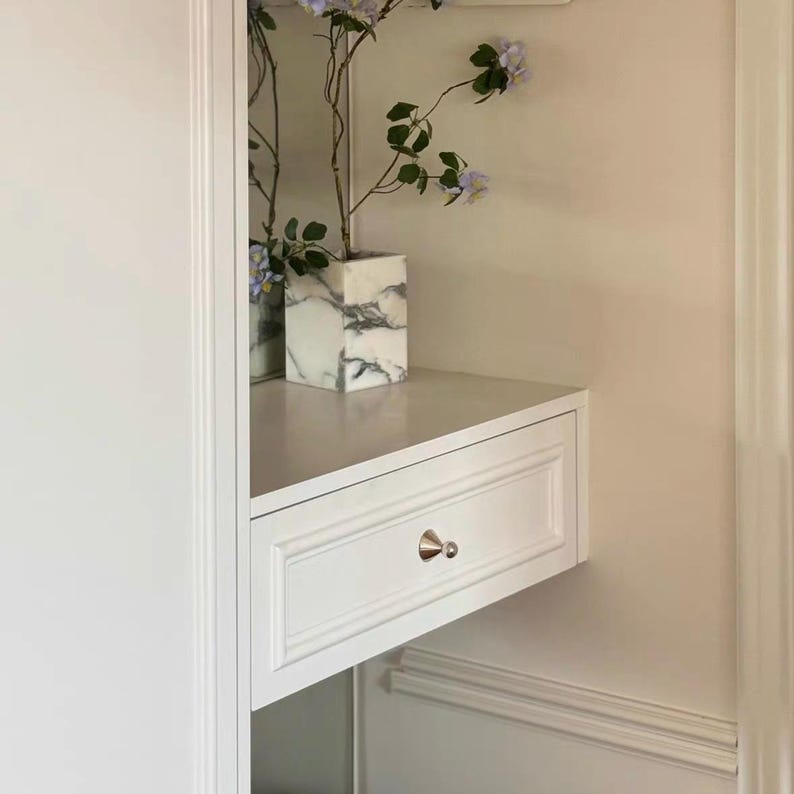 Puede incluir: Un caj&oacute;n flotante blanco con un solo pomo c&oacute;nico plateado, montado en la pared. Encima hay un jarr&oacute;n cuadrado con un patr&oacute;n de m&aacute;rmol blanco y gris, que contiene una rama delgada con peque&ntilde;as flores de color morado claro y hojas verdes.