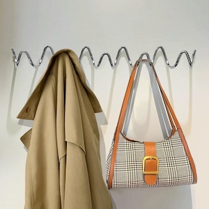 May include: A tan trench coat hangs on a wavy chrome wall hook next to a plaid shoulder bag with brown leather trim and a gold buckle. The bag has a brown leather strap.