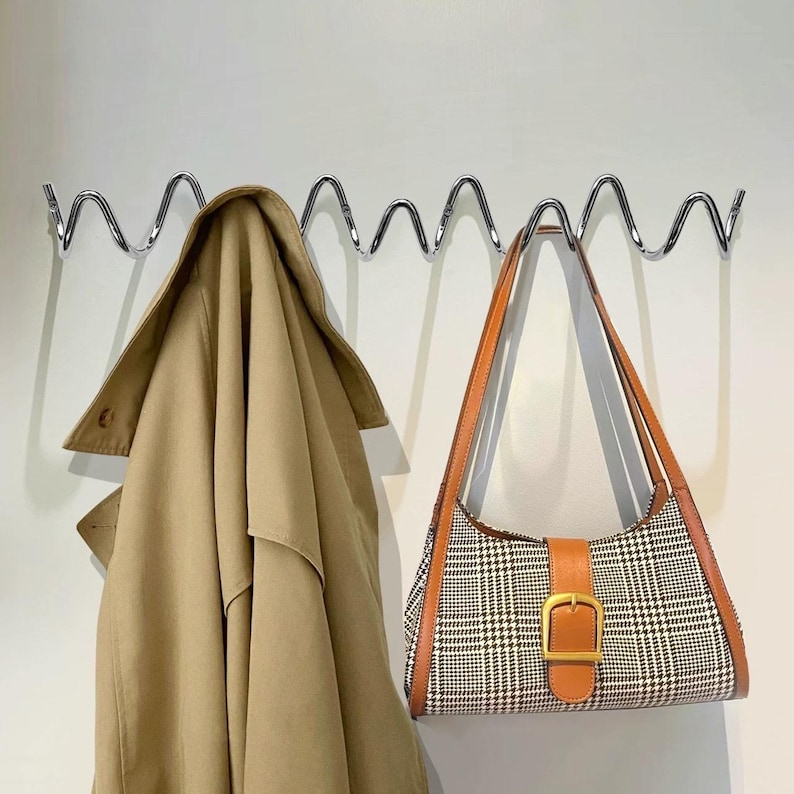 May include: A tan trench coat hangs on a wavy chrome wall hook next to a plaid shoulder bag with brown leather trim and a gold buckle. The bag has a brown leather strap.