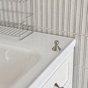 Puede incluir: Un pomo de mueble de metal plateado pulido con base c&oacute;nica y parte superior esf&eacute;rica, montado en un caj&oacute;n de tocador blanco, con un pomo a juego colocado sobre la encimera blanca junto a un estante de alambre y azulejos de pared grises.