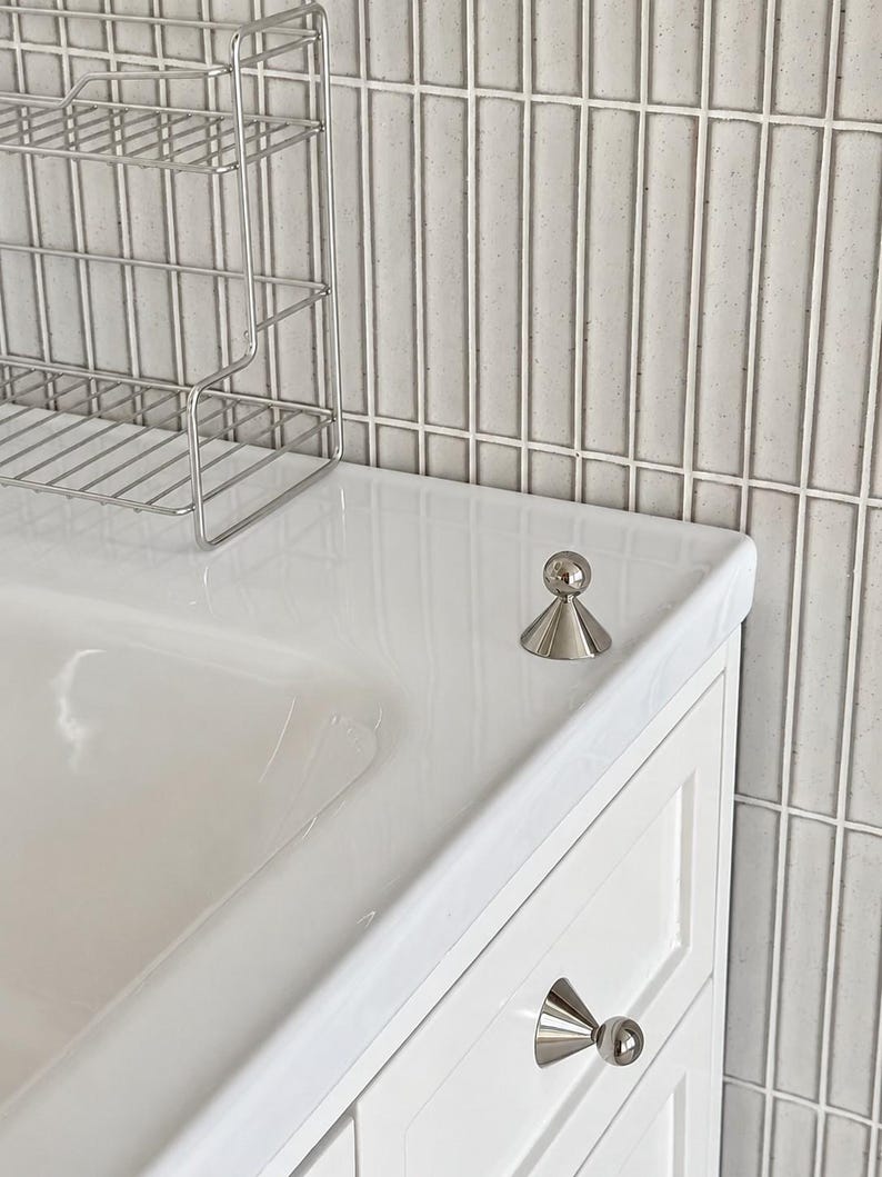 Puede incluir: Un pomo de mueble de metal plateado pulido con base c&oacute;nica y parte superior esf&eacute;rica, montado en un caj&oacute;n de tocador blanco, con un pomo a juego colocado sobre la encimera blanca junto a un estante de alambre y azulejos de pared grises.