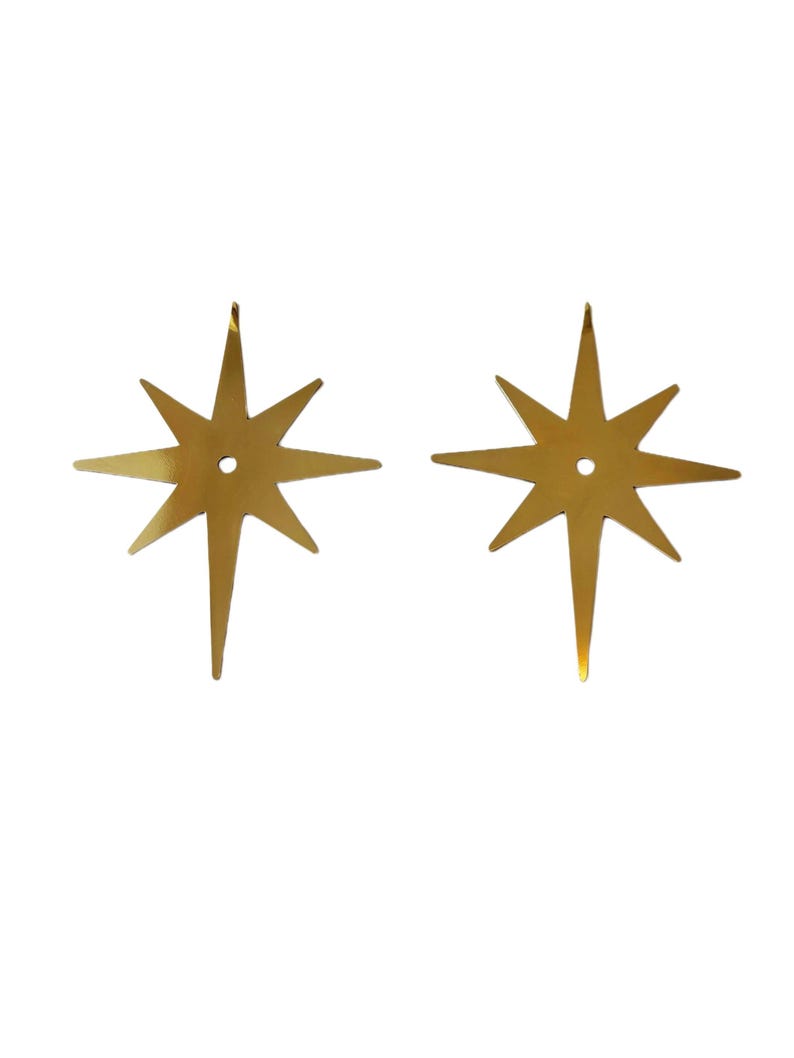 May include: Two identical gold-colored metal star ornaments, each featuring eight points of varying lengths and a small circular hole centered in the middle. The stars have a smooth, reflective surface and a flat, symmetrical design.