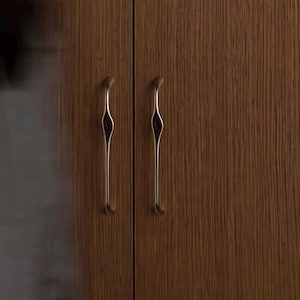 May include: Two vertical, slender, metallic cabinet handles with a smooth, curved, and tapered design. The handles are mounted on a dark brown wooden cabinet door surface that features a prominent, vertical wood grain texture throughout.