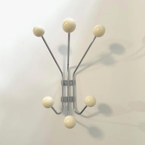 May include: A wall-mounted coat rack with a chrome finish and six cream-colored ball-shaped hooks. The hooks extend from a central metal support, creating a modern and functional design. The rack is mounted on a white wall.