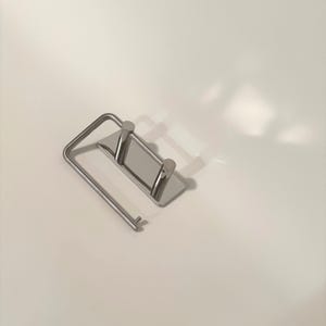 May include: A silver-colored metal toilet paper holder featuring a rectangular wall-mounting plate with two vertical cylindrical posts and a hinged, U-shaped arm that extends outward to hold a roll of paper in a horizontal position.