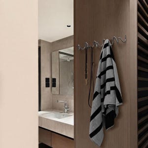 May include: A chrome metal wall-mounted coat rack with a wavy design. A black and gray striped towel and a black leash hang from the hooks. The rack is mounted on a wooden wall in a bathroom setting.
