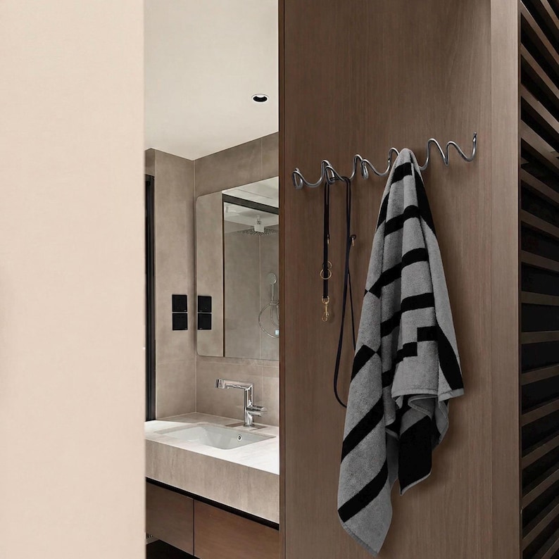 May include: A chrome metal wall-mounted coat rack with a wavy design. A black and gray striped towel and a black leash hang from the hooks. The rack is mounted on a wooden wall in a bathroom setting.