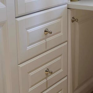 Puede incluir: Un conjunto de armarios de madera blanca con tres cajones apilados y una puerta adyacente, cada uno equipado con un pomo c&oacute;nico de tono plateado pulido con punta esf&eacute;rica, ofreciendo una soluci&oacute;n de almacenamiento funcional.