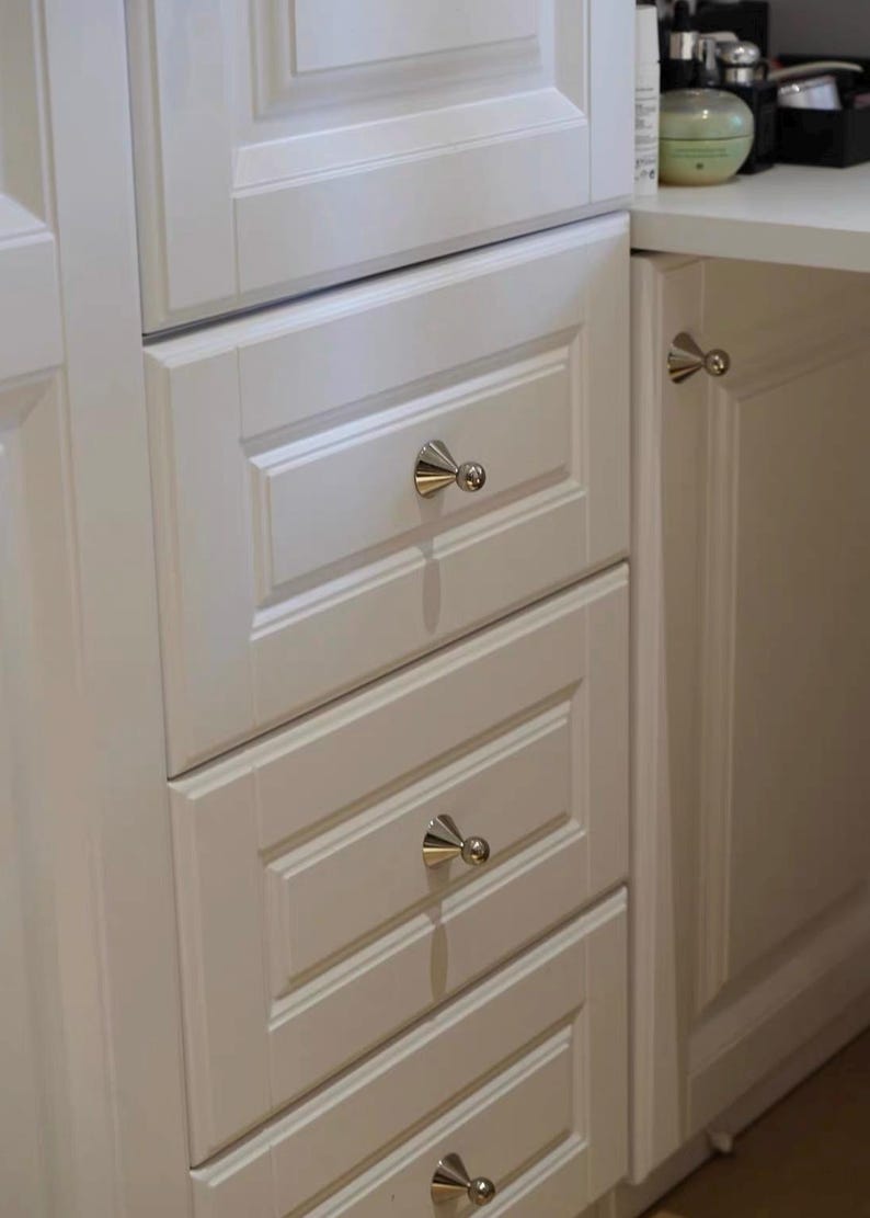 Puede incluir: Un conjunto de armarios de madera blanca con tres cajones apilados y una puerta adyacente, cada uno equipado con un pomo c&oacute;nico de tono plateado pulido con punta esf&eacute;rica, ofreciendo una soluci&oacute;n de almacenamiento funcional.
