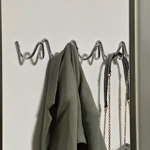 May include: A chrome metal wall-mounted coat rack with a wavy design. A green jacket and a gray purse with a gold chain are hanging on the rack. The rack is mounted on a white wall.
