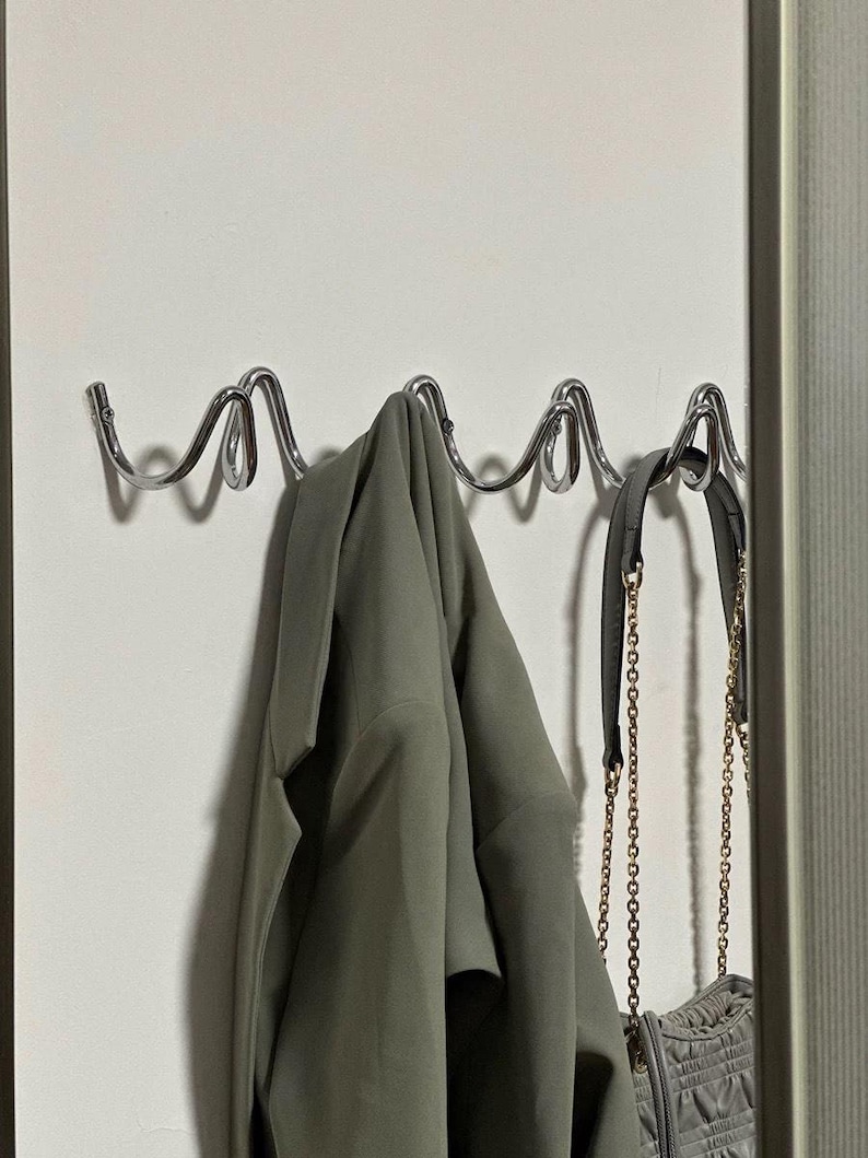 May include: A chrome metal wall-mounted coat rack with a wavy design. A green jacket and a gray purse with a gold chain are hanging on the rack. The rack is mounted on a white wall.