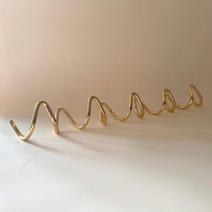 May include: A gold-toned metal wall hook rack featuring a continuous, undulating wave-like design. The piece consists of a single, polished metallic rod bent into a series of repeating, rounded loops that serve as individual hooks for hanging items.