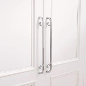 May include: Two long, vertical, polished chrome cabinet handles mounted on white double doors. Each handle features a straight cylindrical bar with decorative, curved scroll-like ends at the top and bottom, providing a sleek, metallic contrast.