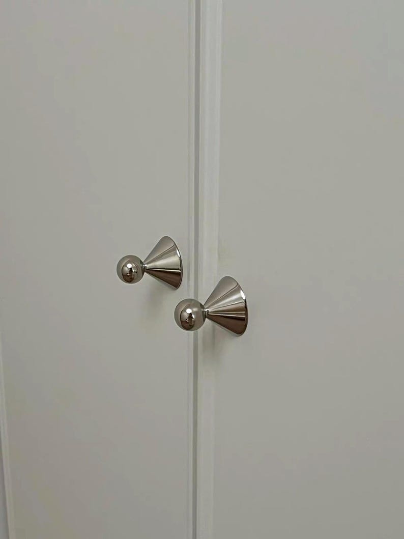 Puede incluir: Dos pomos de armario de metal plateado pulido con base c&oacute;nica y parte superior esf&eacute;rica, montados sobre una superficie de puerta de armario blanca lisa. Los herrajes presentan un acabado liso y reflectante y una forma geom&eacute;trica.