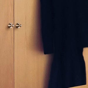 Puede incluir: Un armario de madera de color claro con dos puertas verticales que tienen peque&ntilde;os pomos met&aacute;licos redondos de color plateado. Una prenda de color oscuro cuelga parcialmente visible a la derecha del armario, junto a una estanter&iacute;a de madera clara.