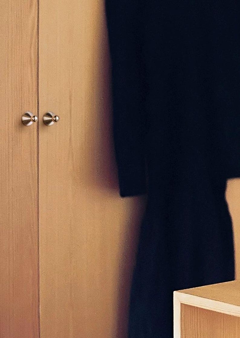 Puede incluir: Un armario de madera de color claro con dos puertas verticales que tienen peque&ntilde;os pomos met&aacute;licos redondos de color plateado. Una prenda de color oscuro cuelga parcialmente visible a la derecha del armario, junto a una estanter&iacute;a de madera clara.