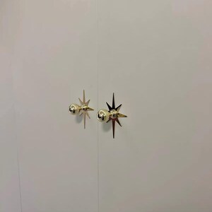 May include: Two gold-colored metal cabinet knobs mounted on a plain white surface. Each knob features a spherical head attached to a flat, multi-pointed star-shaped backplate with sharp, elongated rays extending outward in a symmetrical pattern.