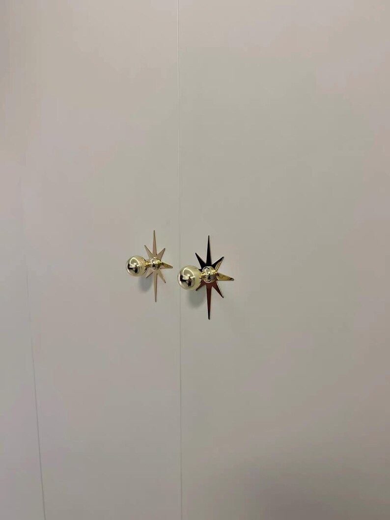 May include: Two gold-colored metal cabinet knobs mounted on a plain white surface. Each knob features a spherical head attached to a flat, multi-pointed star-shaped backplate with sharp, elongated rays extending outward in a symmetrical pattern.