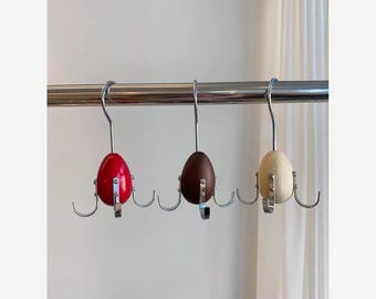 Puede incluir: Tres perchas con forma de huevo en rojo, marrón y crema. Cada percha tiene un gancho metálico y cuatro ganchos más pequeños debajo. Las perchas están suspendidas de una barra metálica.