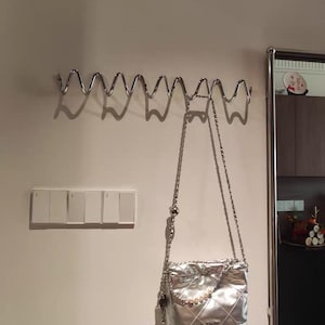 May include: A silver quilted handbag with a chain strap hangs from a wavy metal wall hook. The bag has a pearl accent and a diamond pattern. A set of light switches is visible on the wall.