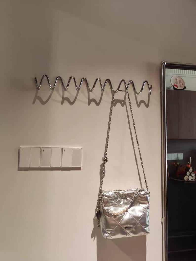 May include: A silver quilted handbag with a chain strap hangs from a wavy metal wall hook. The bag has a pearl accent and a diamond pattern. A set of light switches is visible on the wall.