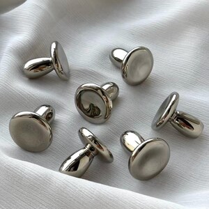 Puede incluir: Un conjunto de seis botones de metal en tono plateado con un dise&ntilde;o liso, reflectante y en forma de seta. Cada bot&oacute;n presenta una tapa redondeada y pulida con una base de v&aacute;stago resistente, mostrando un acabado met&aacute;lico uniforme.