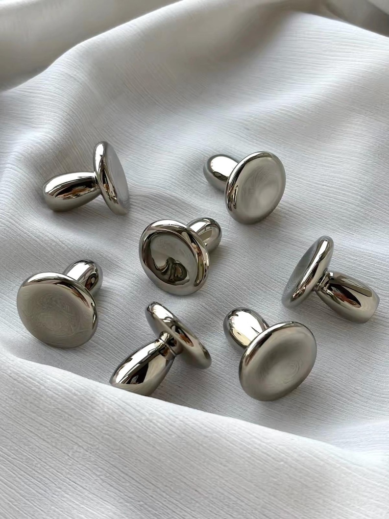 Puede incluir: Un conjunto de seis botones de metal en tono plateado con un dise&ntilde;o liso, reflectante y en forma de seta. Cada bot&oacute;n presenta una tapa redondeada y pulida con una base de v&aacute;stago resistente, mostrando un acabado met&aacute;lico uniforme.