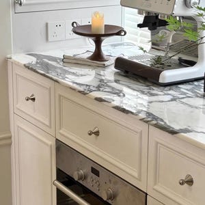 Puede incluir: Un mueble de cocina blanco con encimera de m&aacute;rmol, que incluye un horno de acero inoxidable con mandos de control, una cafetera espresso blanca y un cuenco de pedestal marr&oacute;n oscuro con una vela de pilar blanca sobre un pa&ntilde;o claro plegado.