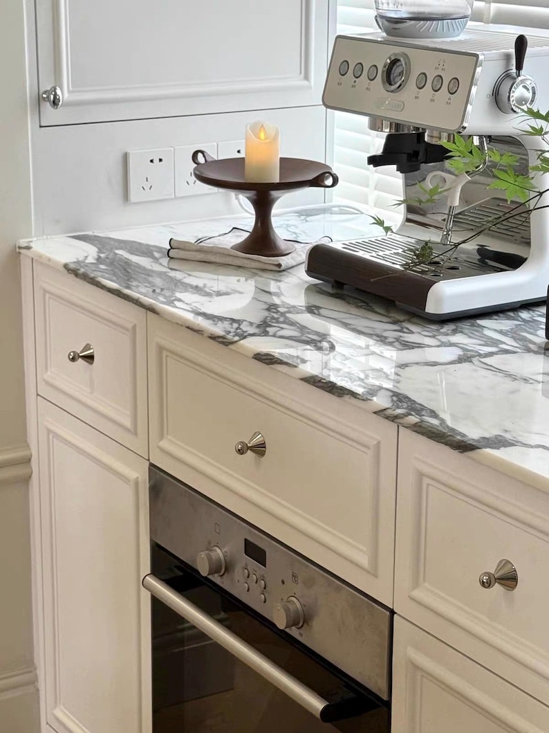 Puede incluir: Un mueble de cocina blanco con encimera de m&aacute;rmol, que incluye un horno de acero inoxidable con mandos de control, una cafetera espresso blanca y un cuenco de pedestal marr&oacute;n oscuro con una vela de pilar blanca sobre un pa&ntilde;o claro plegado.