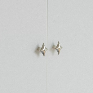 May include: Two polished silver-toned cabinet knobs with a four-pointed star shape attached to a light gray, flat-surfaced double door. The hardware features a smooth, reflective metallic finish and a symmetrical, geometric design.