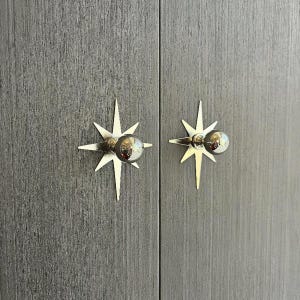 May include: Two silver-toned starburst-shaped cabinet knobs mounted on a dark gray wood-grain textured surface. Each knob features a central spherical handle attached to a flat, multi-pointed star backplate with a metallic finish.