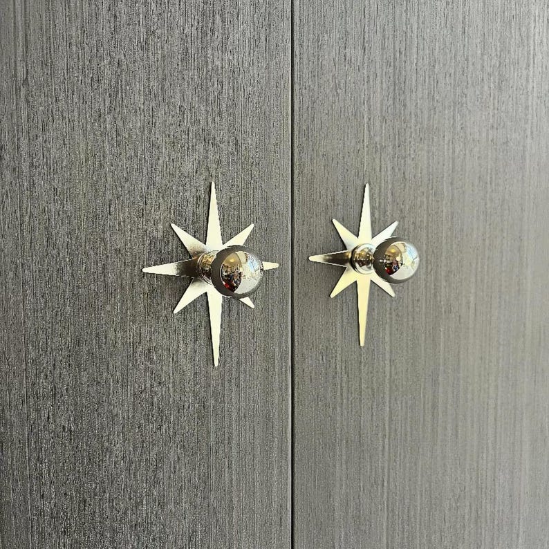 May include: Two silver-toned starburst-shaped cabinet knobs mounted on a dark gray wood-grain textured surface. Each knob features a central spherical handle attached to a flat, multi-pointed star backplate with a metallic finish.