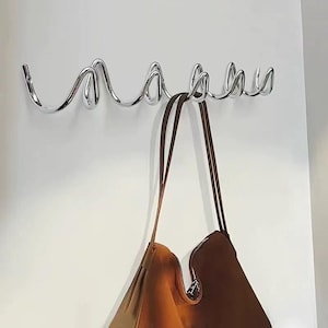 May include: A chrome metal wall-mounted coat rack with a wavy design. A brown leather tote bag hangs from the hooks. The rack has six curved hooks and is mounted on a white wall.