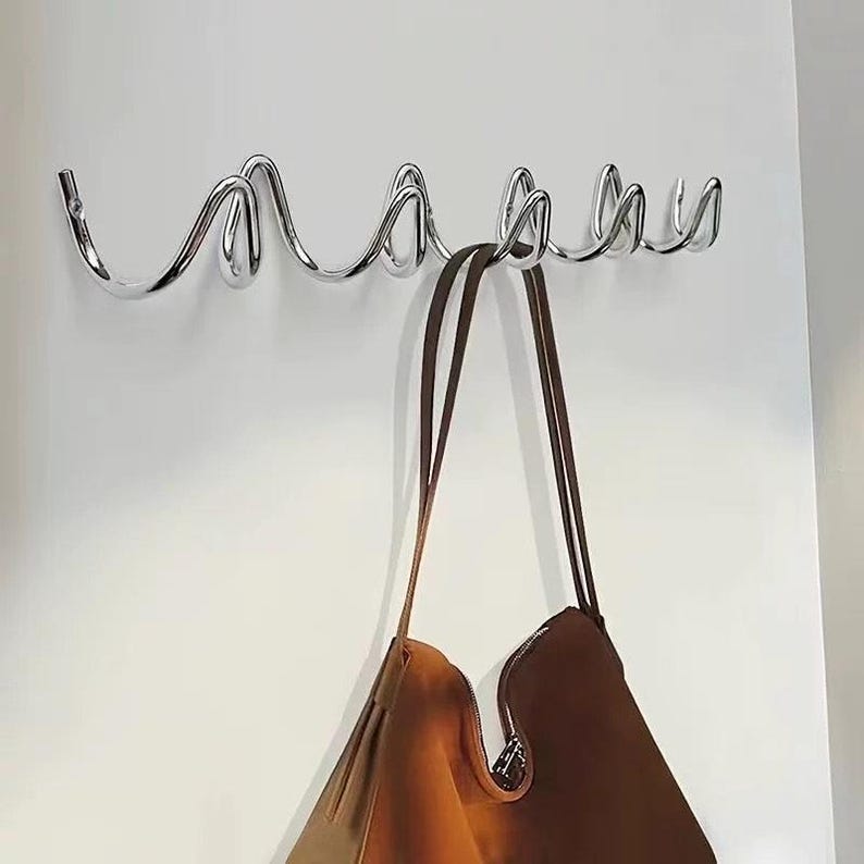 May include: A chrome metal wall-mounted coat rack with a wavy design. A brown leather tote bag hangs from the hooks. The rack has six curved hooks and is mounted on a white wall.