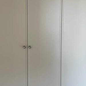 Puede incluir: Un conjunto de puertas de armario blancas con un acabado mate liso y paneles verticales. Cada puerta est&aacute; equipada con un peque&ntilde;o pomo de metal plateado pulido de forma circular, centrado horizontalmente para facilitar el acceso y la est&eacute;tica.