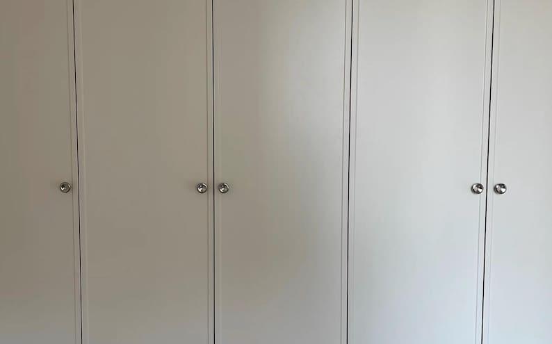 Puede incluir: Un conjunto de puertas de armario blancas con un acabado mate liso y paneles verticales. Cada puerta est&aacute; equipada con un peque&ntilde;o pomo de metal plateado pulido de forma circular, centrado horizontalmente para facilitar el acceso y la est&eacute;tica.