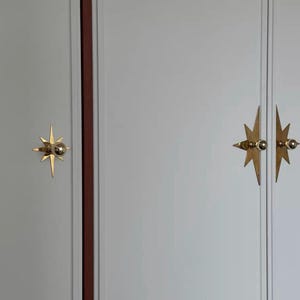 K&ouml;nnte beinhalten: Ein hellgrauer Schrank mit drei goldfarbenen, sternf&ouml;rmigen Metallgriffen. Die Griffe bestehen aus einem zentralen kugelf&ouml;rmigen Knopf auf einer spitzen, mehrstrahligen Sternplatte. Die Schrankt&uuml;ren weisen vertikale Paneeldetails auf.