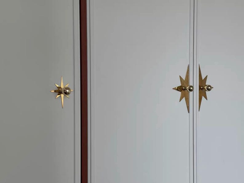 K&ouml;nnte beinhalten: Ein hellgrauer Schrank mit drei goldfarbenen, sternf&ouml;rmigen Metallgriffen. Die Griffe bestehen aus einem zentralen kugelf&ouml;rmigen Knopf auf einer spitzen, mehrstrahligen Sternplatte. Die Schrankt&uuml;ren weisen vertikale Paneeldetails auf.