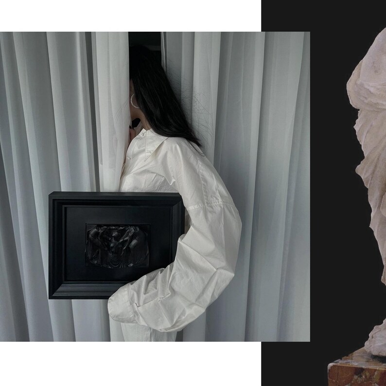 K&ouml;nnte beinhalten: Eine schwarz gerahmte Reliefskulptur wird vor einem wei&szlig;en Vorhang gehalten. Das Relief scheint eine dunkle, monochrome Darstellung von Figuren zu sein. Die Person, die den Rahmen h&auml;lt, tr&auml;gt ein wei&szlig;es Hemd.