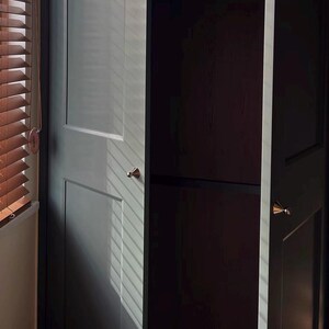 Puede incluir: Un armario de color gris carb&oacute;n oscuro con dos puertas que presentan paneles empotrados y peque&ntilde;os pomos met&aacute;licos c&oacute;nicos. El armario est&aacute; situado junto a una ventana con persianas horizontales marrones que proyectan sombras.