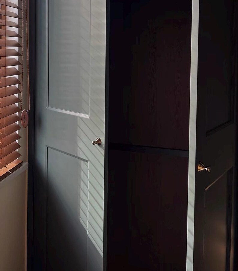 Puede incluir: Un armario de color gris carb&oacute;n oscuro con dos puertas que presentan paneles empotrados y peque&ntilde;os pomos met&aacute;licos c&oacute;nicos. El armario est&aacute; situado junto a una ventana con persianas horizontales marrones que proyectan sombras.