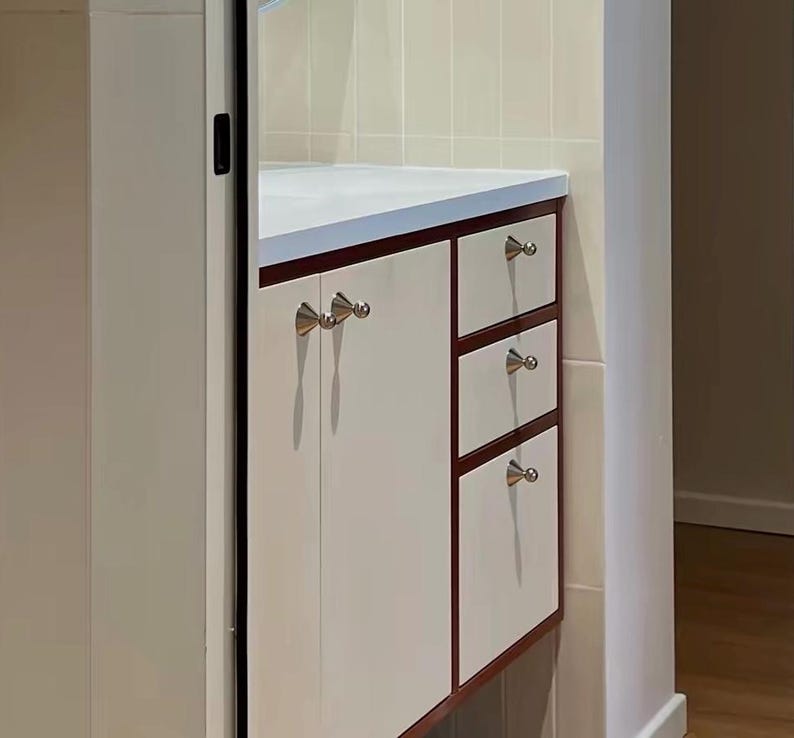 Puede incluir: Un mueble de cocina con puertas y frentes de caj&oacute;n en color blanco roto, enmarcados por un borde de madera oscura. La unidad incluye una puerta de armario grande y tres cajones apilados, con peque&ntilde;os pomos c&oacute;nicos plateados.