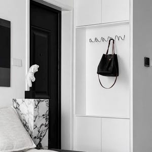 May include: A modern interior with a black door, white walls, and built-in storage. A black leather tote bag hangs from a wavy metal hook. A marble-patterned pedestal holds a white sculpture. A gray sofa and black bench complete the minimalist design.