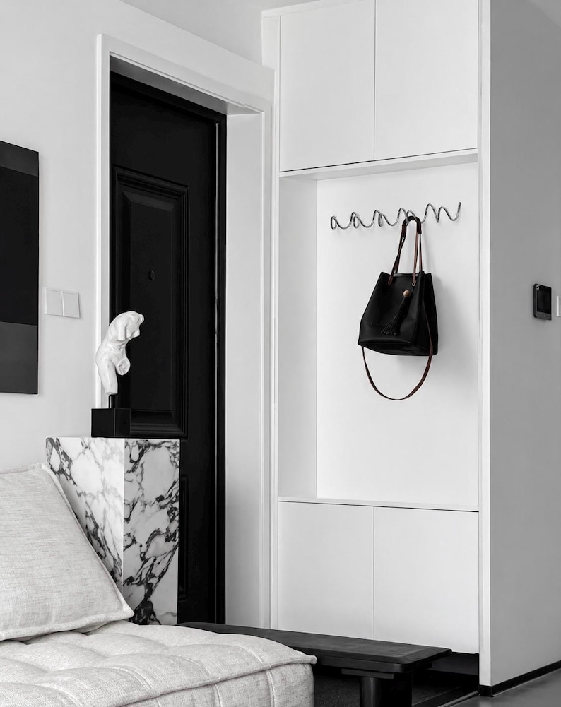 May include: A modern interior with a black door, white walls, and built-in storage. A black leather tote bag hangs from a wavy metal hook. A marble-patterned pedestal holds a white sculpture. A gray sofa and black bench complete the minimalist design.