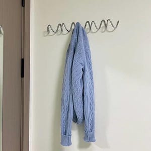 May include: A light blue cable-knit sweater hangs from a silver metal wall hook. The sweater has ribbed cuffs and a classic design. The hook is mounted on a white wall near a closed door.