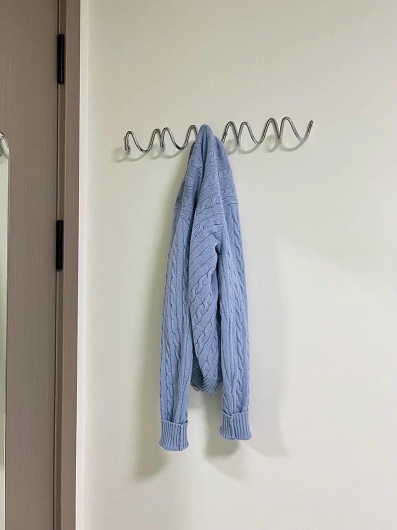 May include: A light blue cable-knit sweater hangs from a silver metal wall hook. The sweater has ribbed cuffs and a classic design. The hook is mounted on a white wall near a closed door.