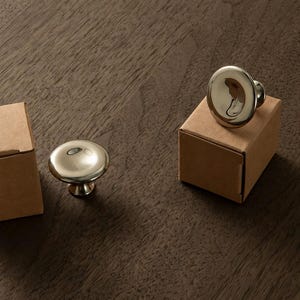 Puede incluir: Dos pomos de armario circulares de metal pulido con una superficie lisa y reflectante. Un pomo descansa sobre una peque&ntilde;a caja de cart&oacute;n cuadrada de color marr&oacute;n claro, mientras que el otro est&aacute; junto a una segunda caja id&eacute;ntica.