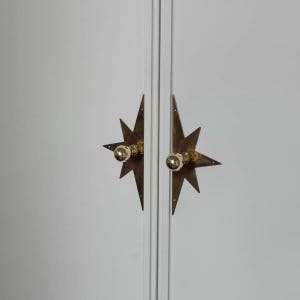 May include: Two brass starburst-shaped cabinet pulls mounted on a white double-door cabinet. Each pull features a central spherical knob attached to a flat, multi-pointed metal backplate with a metallic finish and visible screw holes.